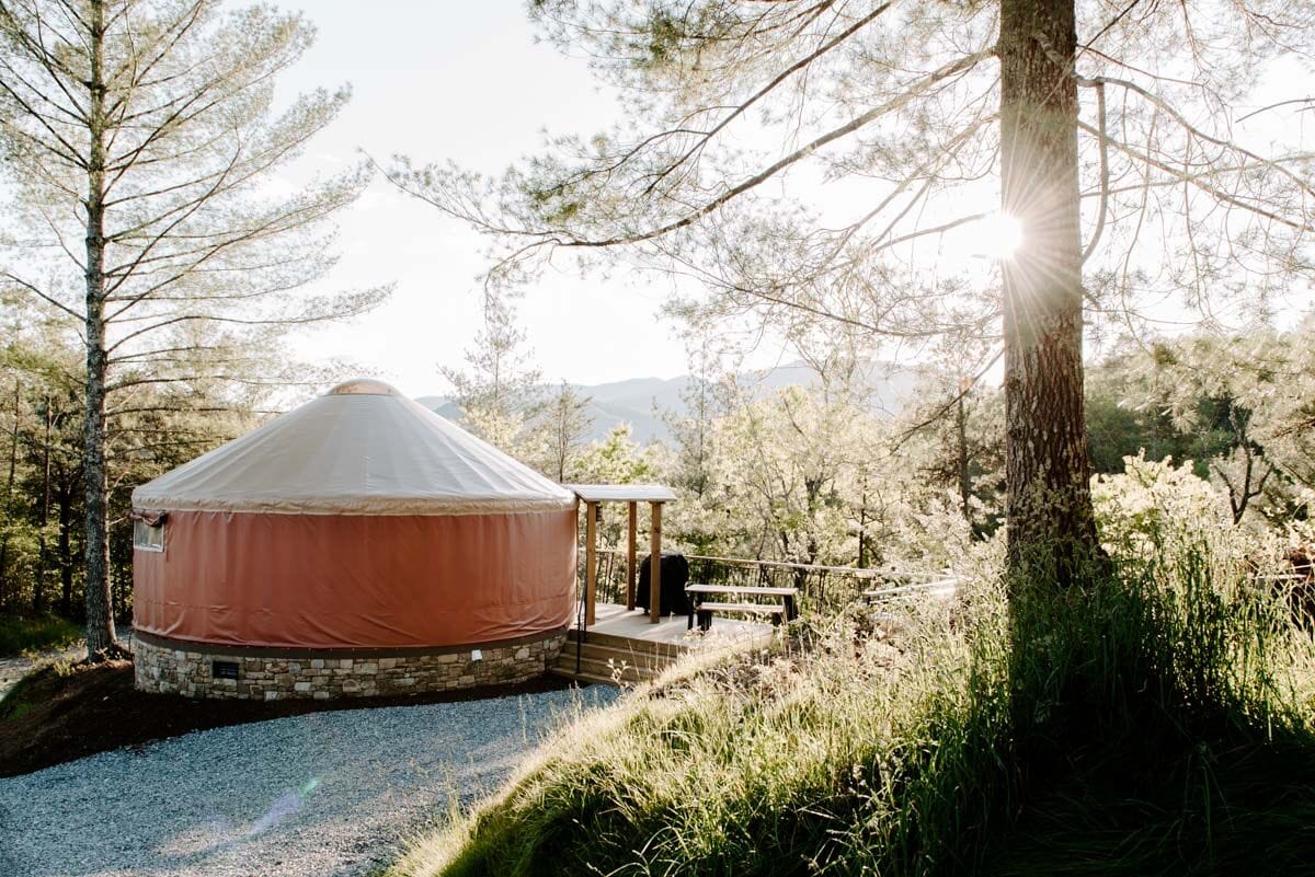 A yurt with a stone foundation, surrounded by trees and a gravel path.