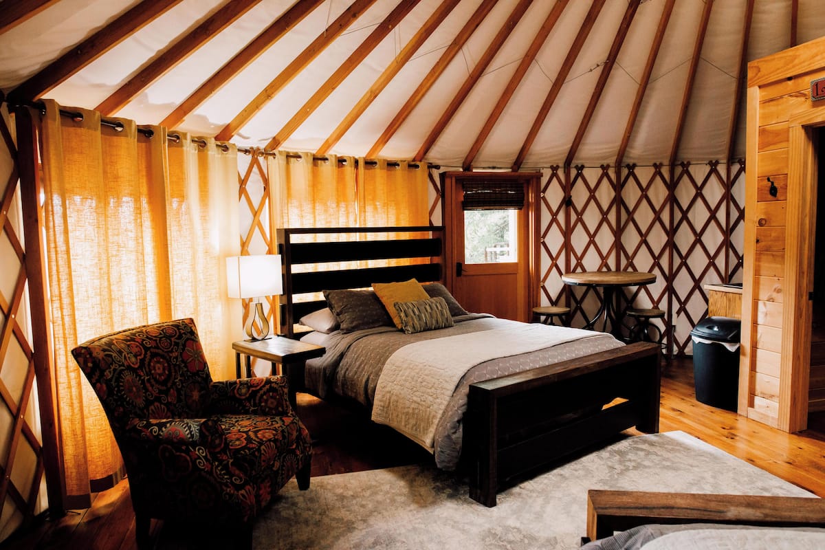 A cozy, well-lit tent-style room with a bed, a chair, a small table, and a window.