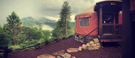 A rustic yurt with a stone staircase leading to a wooden deck, surrounded by a forested landscape.