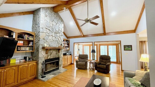A spacious living room with a stone fireplace, wooden beams, a ceiling fan, and a television.