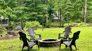 A fire pit surrounded by four Adirondack chairs in a grassy outdoor area with trees and a small building in the background.
