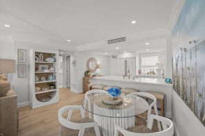 Uma cozinha moderna com armários brancos, uma ilha central e uma área de refeições com mesa de vidro e cadeiras brancas.