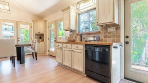 A modern kitchen with light wood cabinets, a black dishwasher, and a window with a view of greenery. - Canyon Treehouse (San Diego)