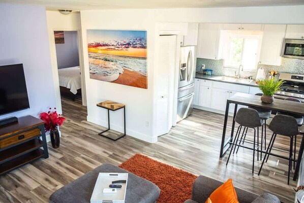 A modern living room with a TV, sofa, and artwork, leading to a kitchen with a dining area.