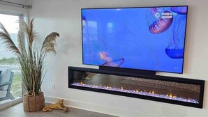 A modern living room with a flat-screen TV mounted on the wall, a built-in fireplace below it, and a potted plant to the left. - Destin Beachfront Oasis (Miramar Beach)