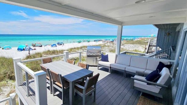 A covered outdoor area with a table and chairs, a sofa, and a grill. It overlooks a beach with umbrellas and people. - Destin Beachfront Oasis (Miramar Beach)