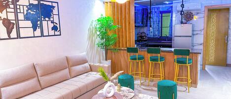 Una sala de estar moderna con un sofá color beige, una mesa de centro redonda y una zona de bar con sillas verdes.