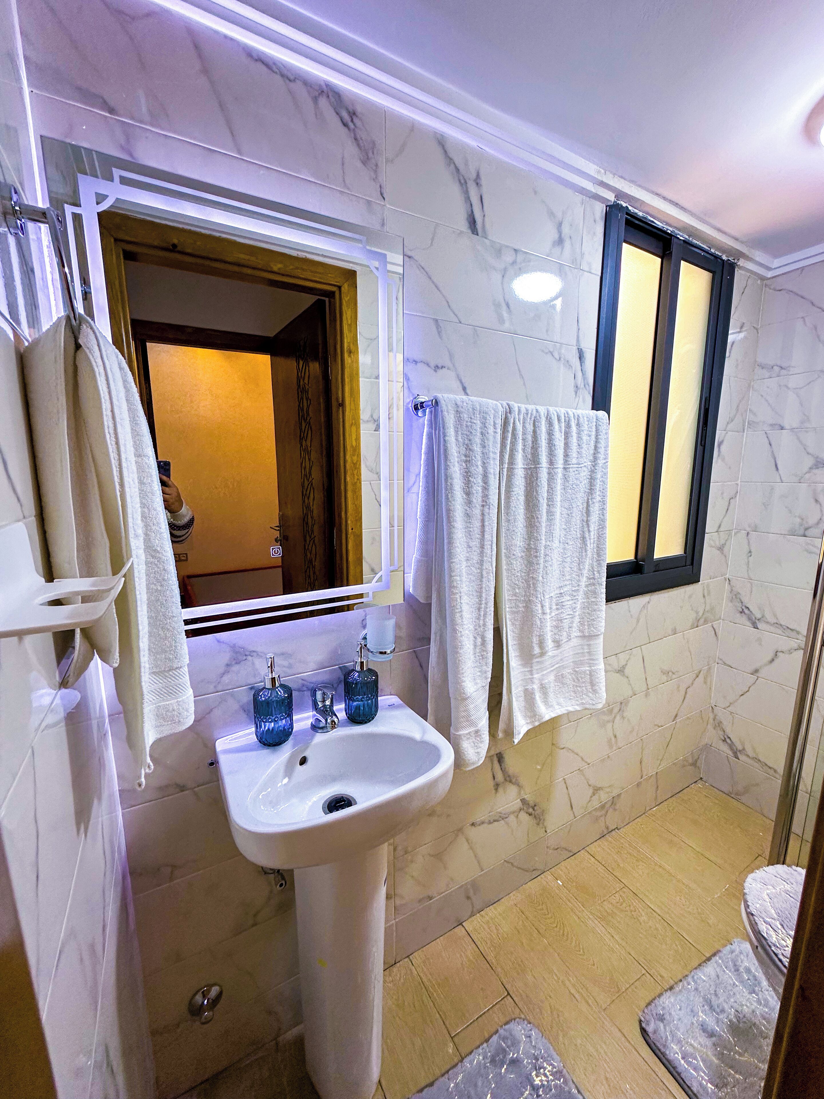 A bathroom with a marble wall, a sink with blue glass soap dispensers, and a window with a towel hanging on the outside.
