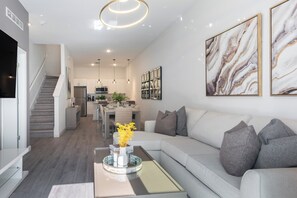 Uma sala de estar moderna com um sofá cinzento, uma mesa de centro com um vaso de flores e uma escadaria que dá para a cozinha.
