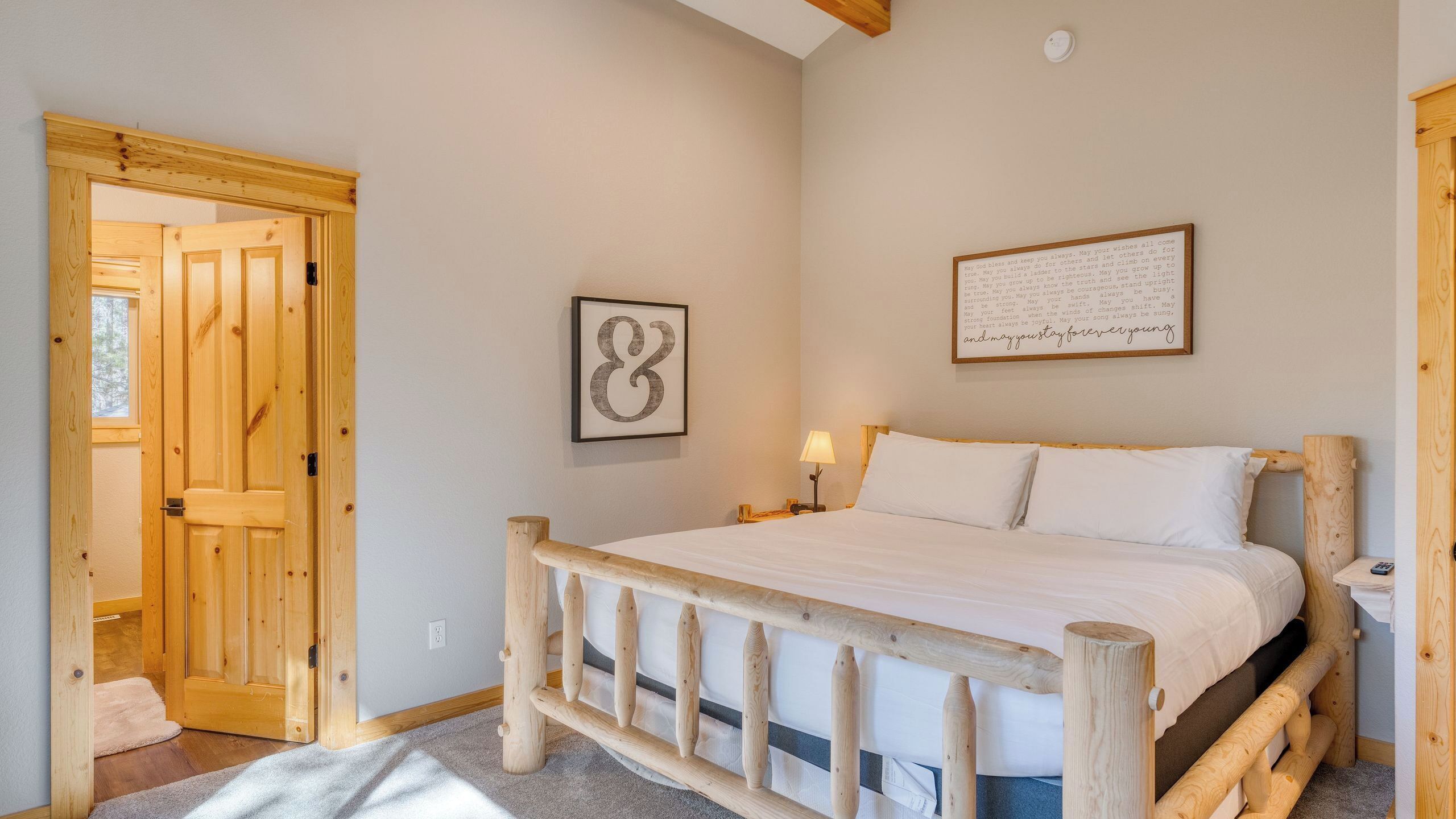A bedroom with a wooden bed frame, a single bed, a nightstand with a lamp, a framed inspirational quote, and a door with a wooden frame.