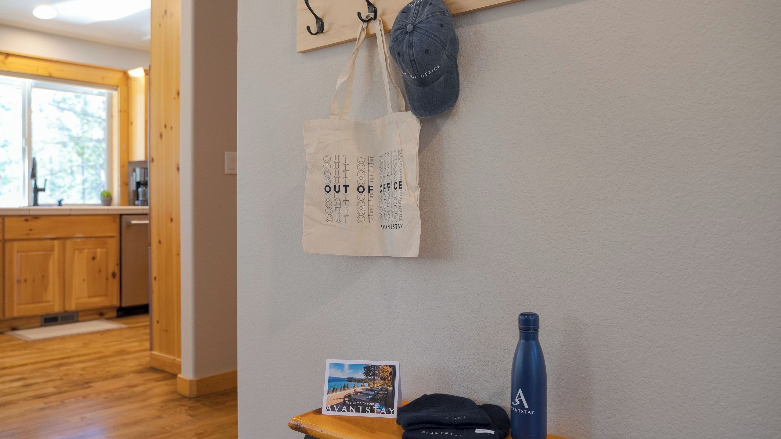 A room with a wooden floor, a kitchen area with wooden cabinets, a hanging bag with text, a blue water bottle, and a welcome sign.