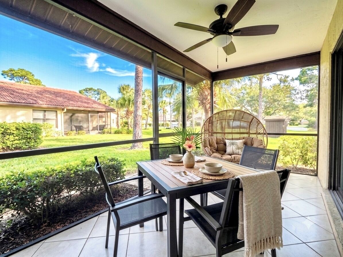 A covered patio with a table set for two, a ceiling fan, and a view of a house and greenery.