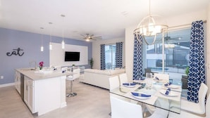 A modern kitchen with a central island, a dining area with a glass table, and a living space with a sofa and a TV.