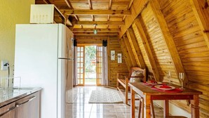 Een keuken met een koelkast, houten kastjes, een eettafel met stoelen en een uitzicht door een glazen deur naar buiten.