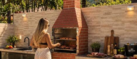A woman grilling outdoors with a brick oven, a stone countertop, and a glass table with a beer.