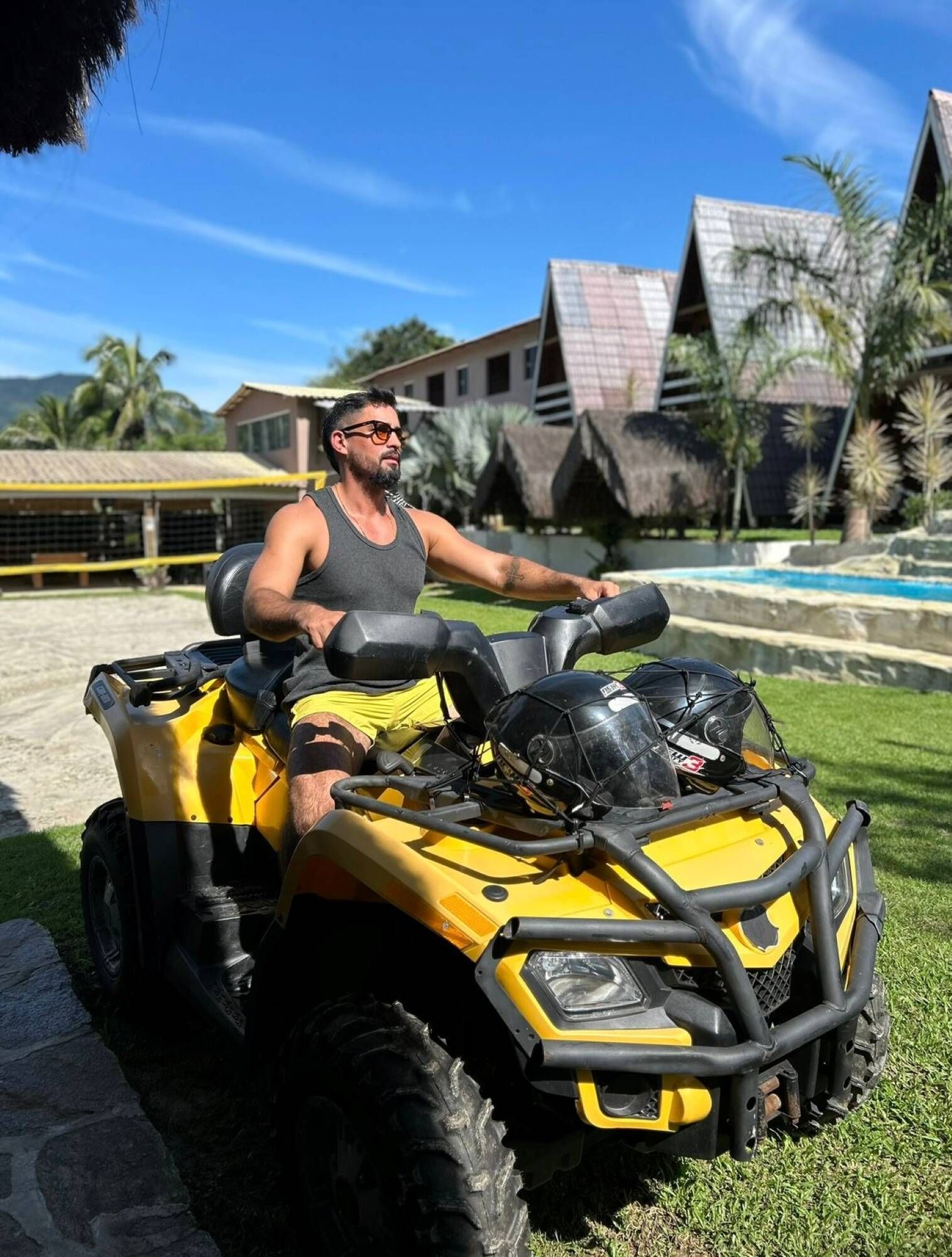Une personne est assise sur un quad jaune dans un espace verdoyant, avec une piscine et des bâtiments en arrière-plan.