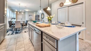 A modern kitchen with a large island, pendant lights, and a dining area with a rug.
