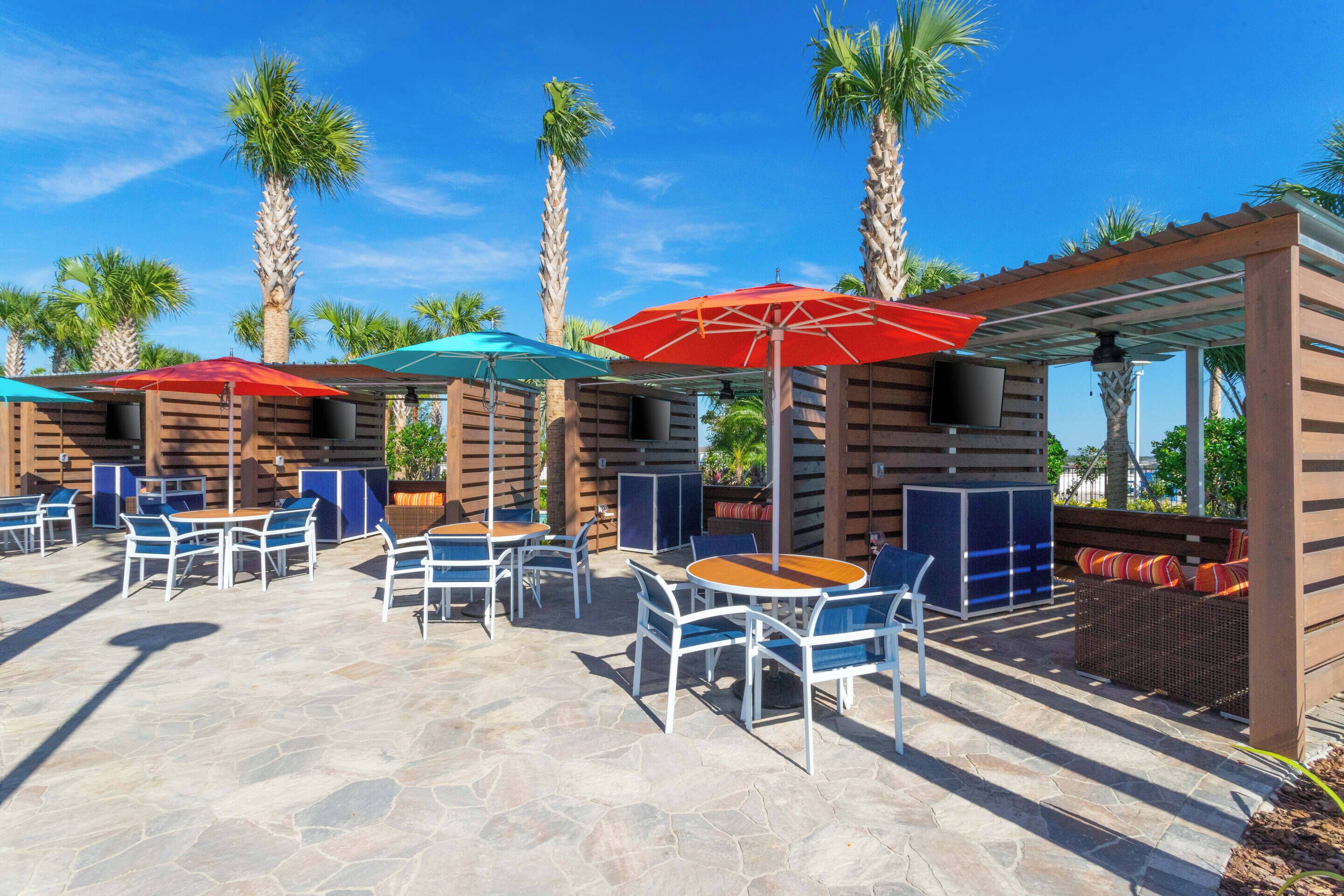 An outdoor patio area with stone flooring, wooden tables and chairs, and umbrellas providing shade.