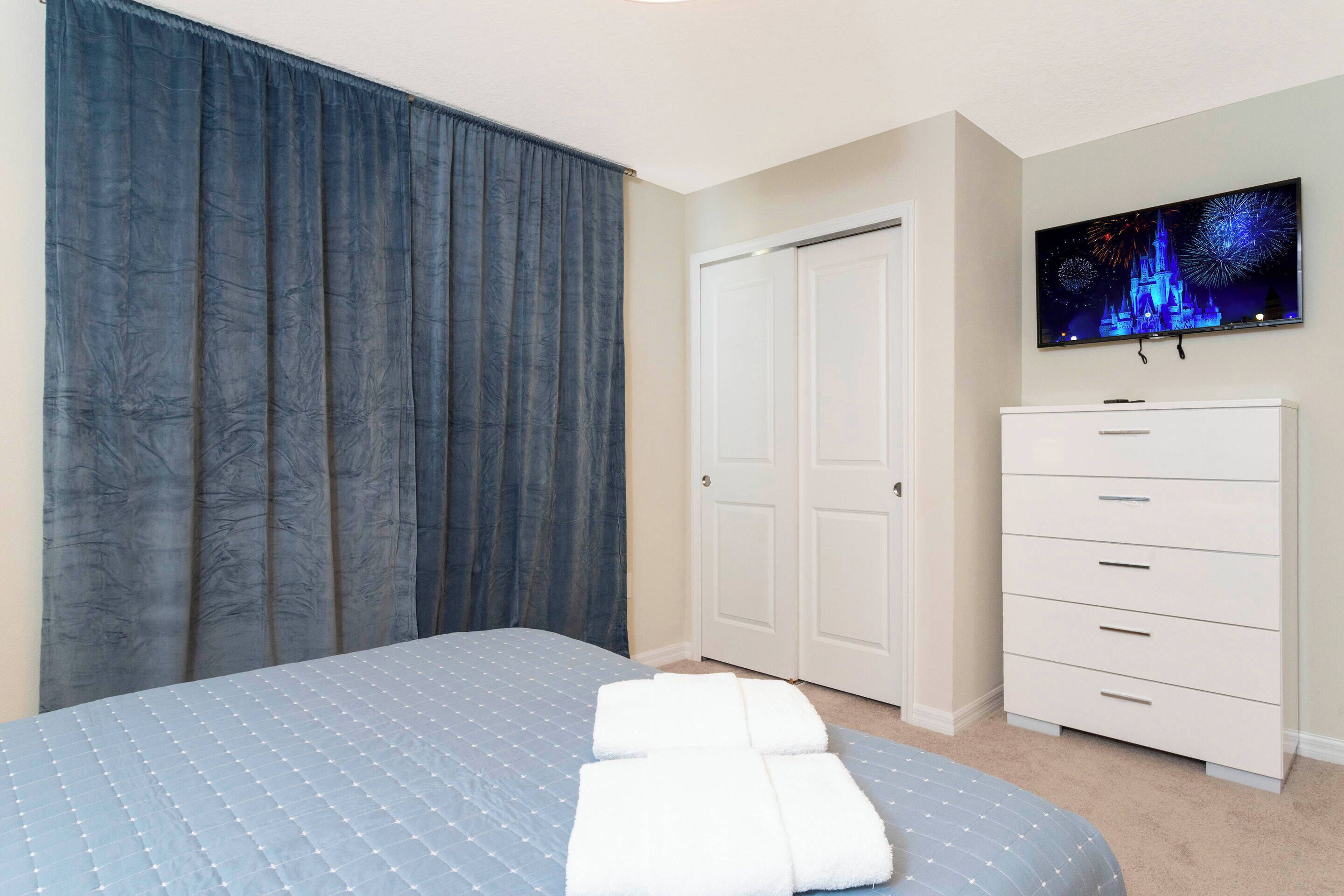 A bedroom with a bed, a wardrobe, a television on the wall, and a large window with curtains.