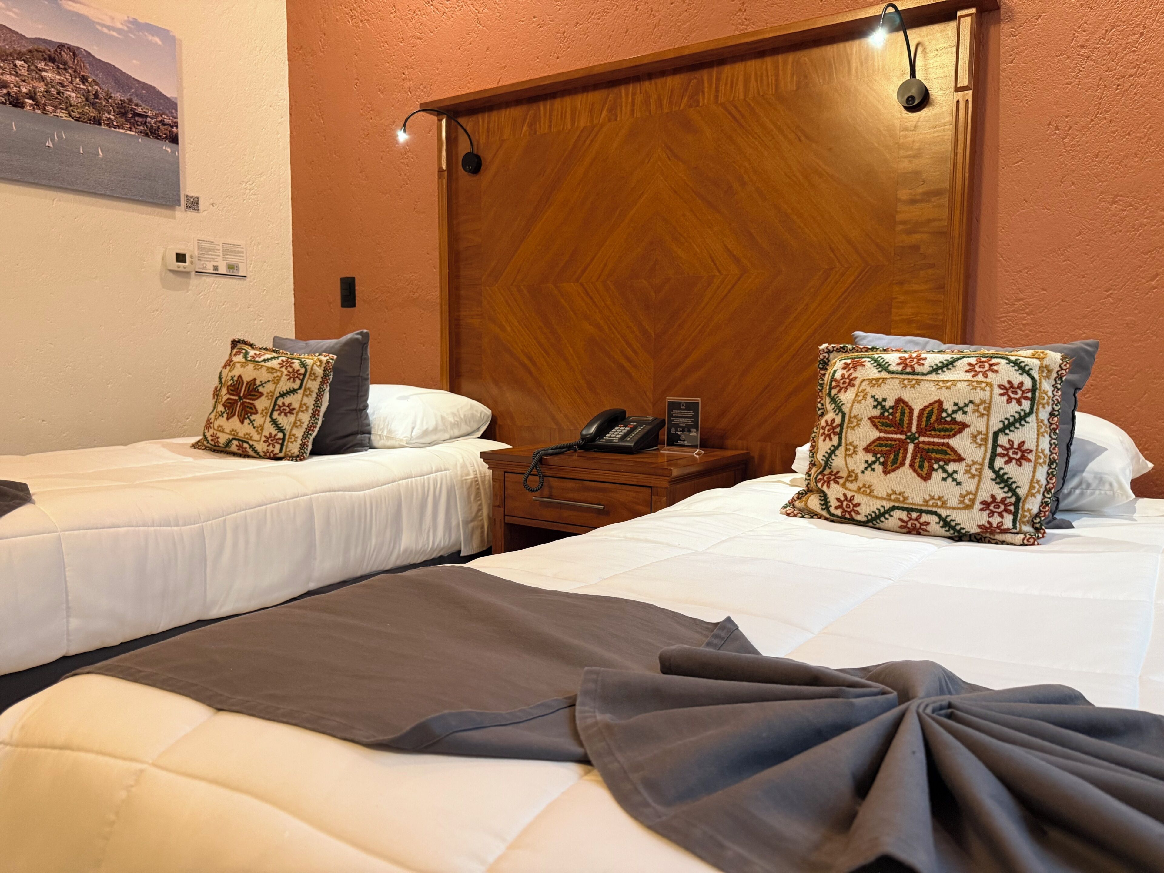 A hotel room with two single beds, a wooden headboard, a bedside table with a phone, and a wall-mounted thermostat.