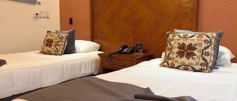 A hotel room with two single beds, a wooden headboard, a bedside table with a phone, and a wall-mounted thermostat.