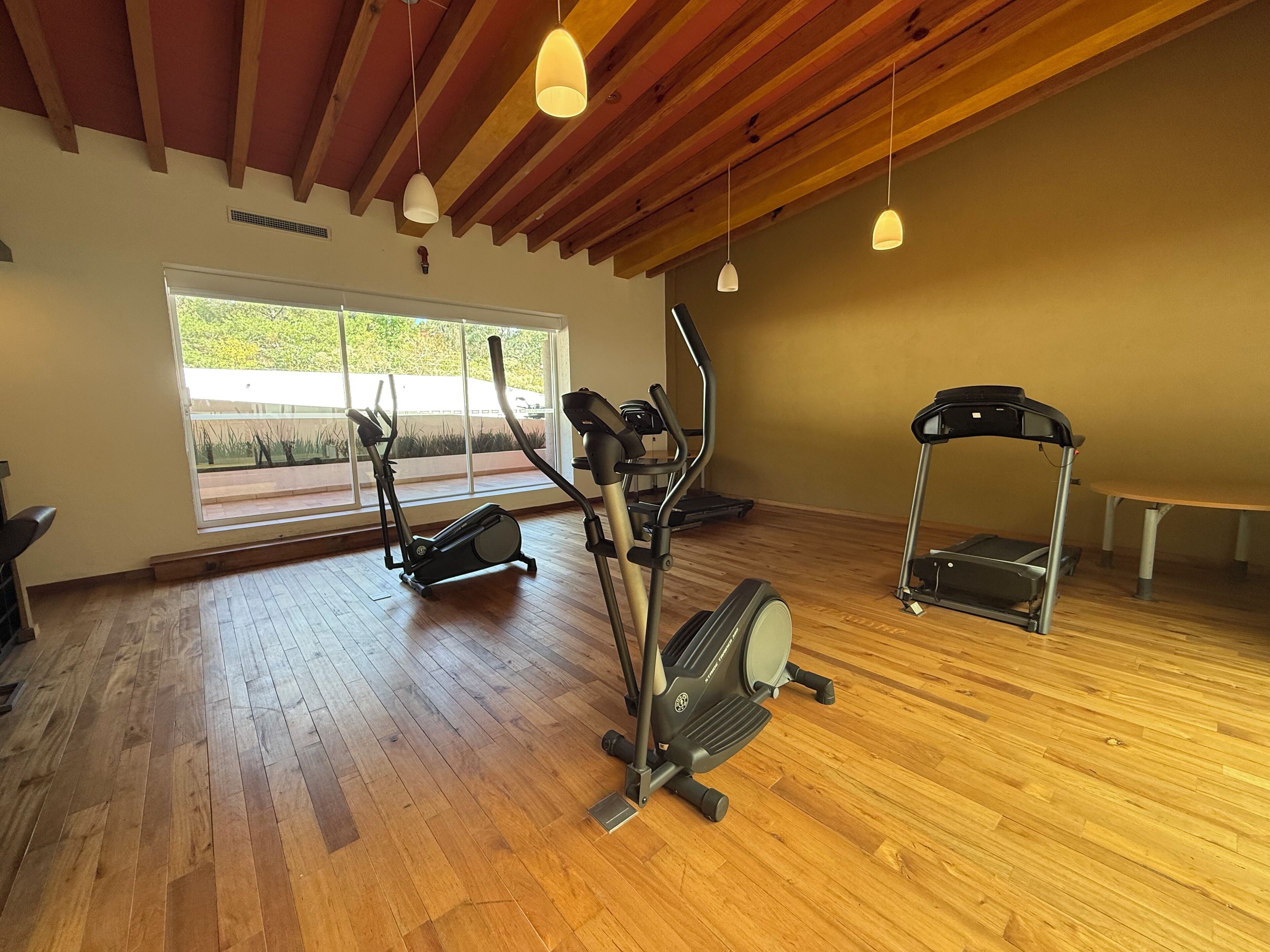 A spacious indoor gym with wooden flooring, exercise equipment, and large windows providing natural light.