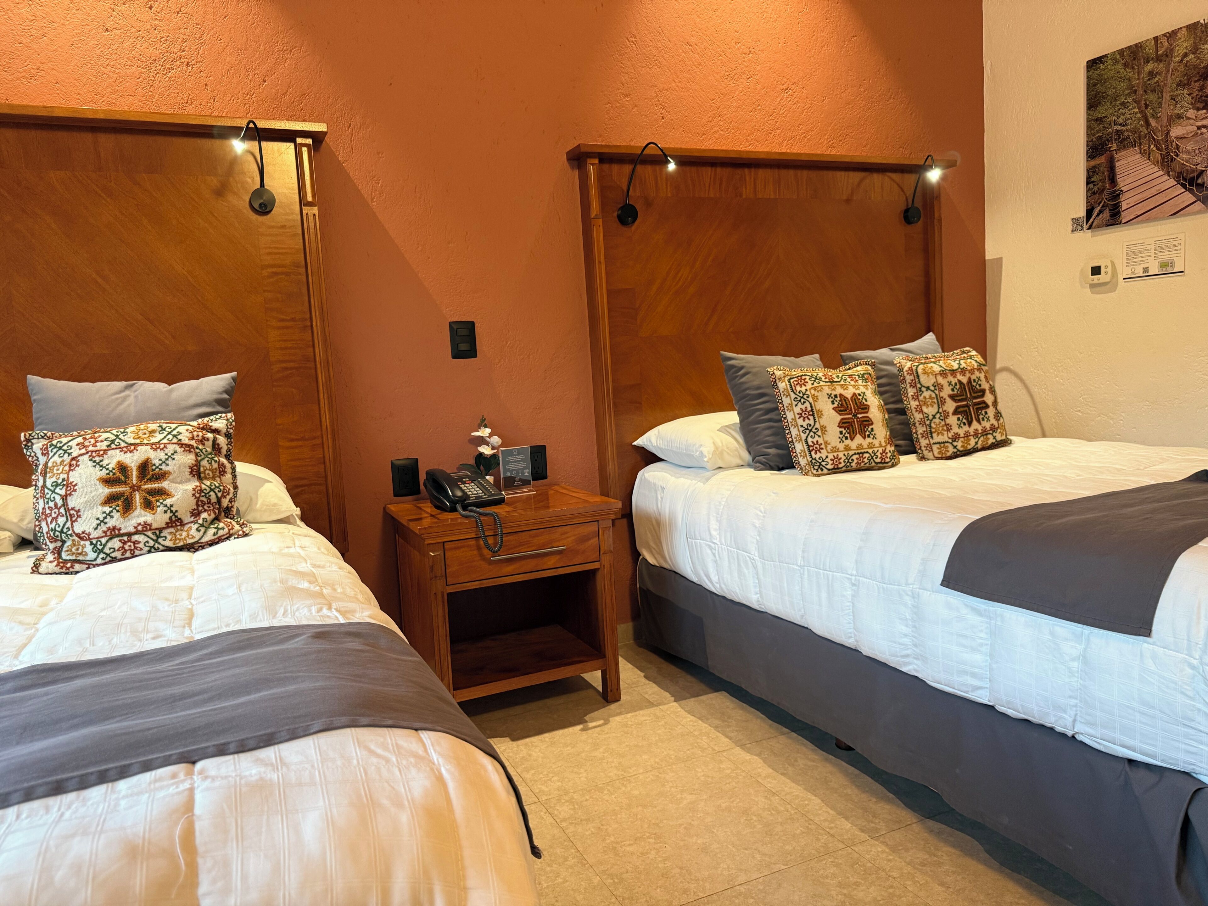 A hotel room with two beds, a wooden nightstand with a telephone, and a wall-mounted light fixture.
