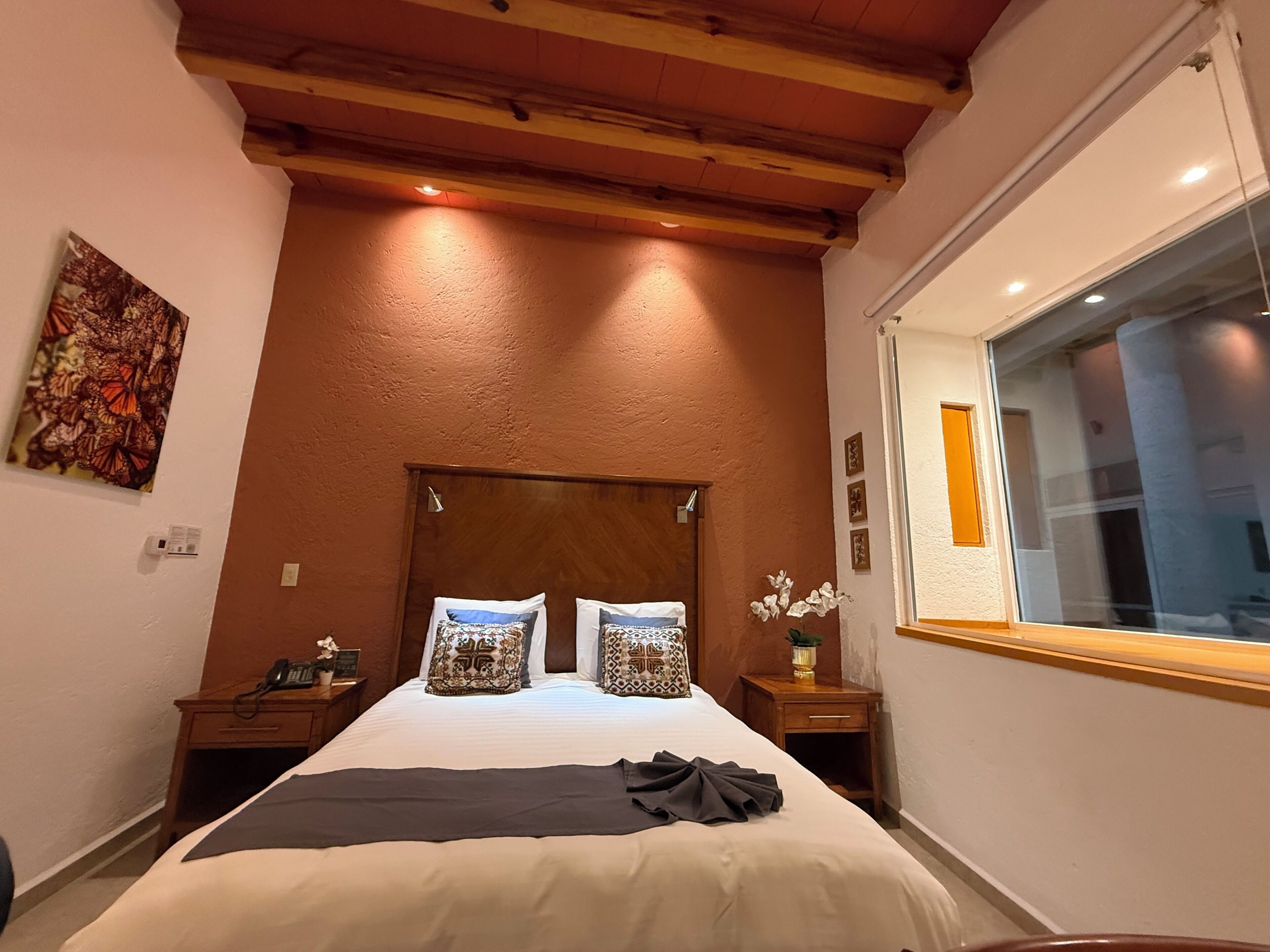 A bedroom with a wooden ceiling, a large bed with patterned pillows, a bedside table with a phone, and a window with a view.