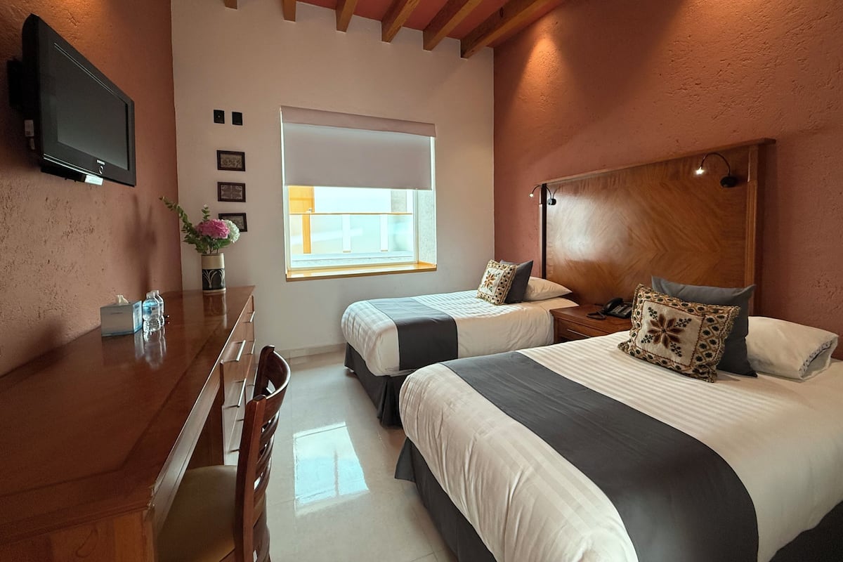 A hotel room with two beds, a desk, a chair, a TV, and a window.