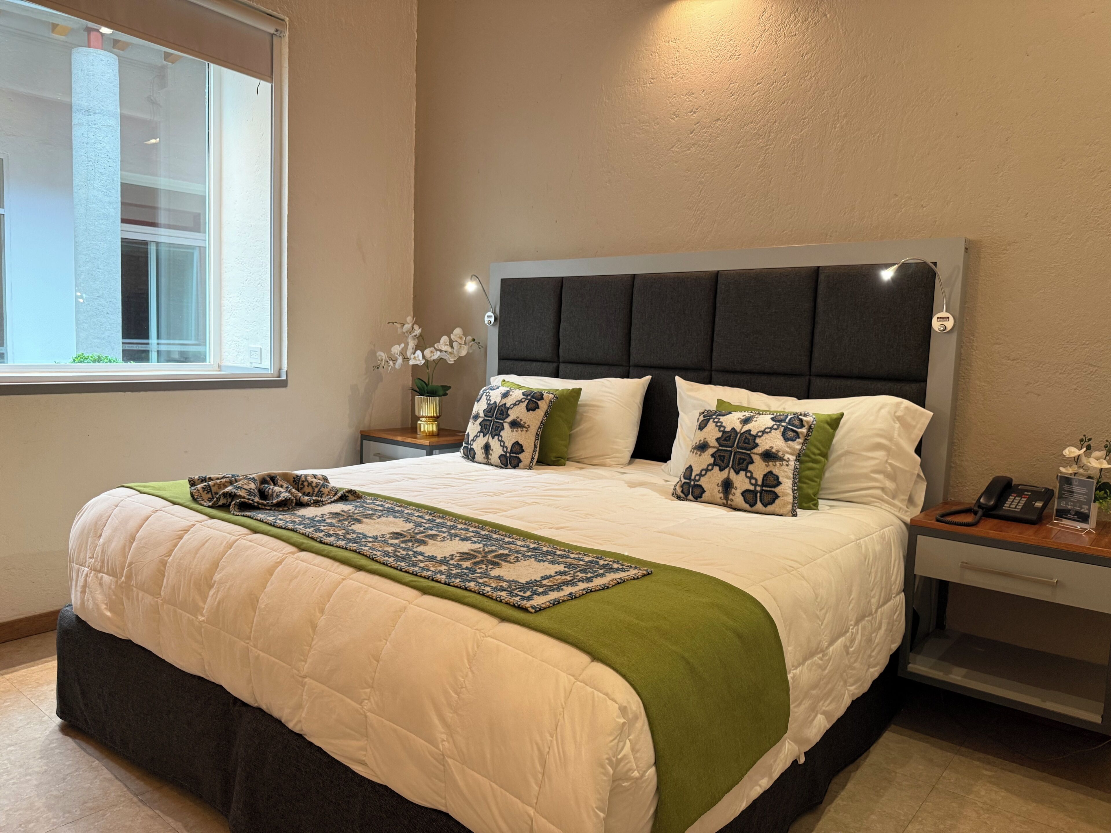 A neatly made bed with a quilt, pillows, and a green throw blanket. There is a bedside table with a lamp and a phone.