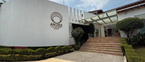 Un edificio dal design moderno, con il logo e la scritta 'ANJIES' sulla facciata. Scale conducono all'ingresso, e un'area giardino curata con siepi potate e un vialetto.