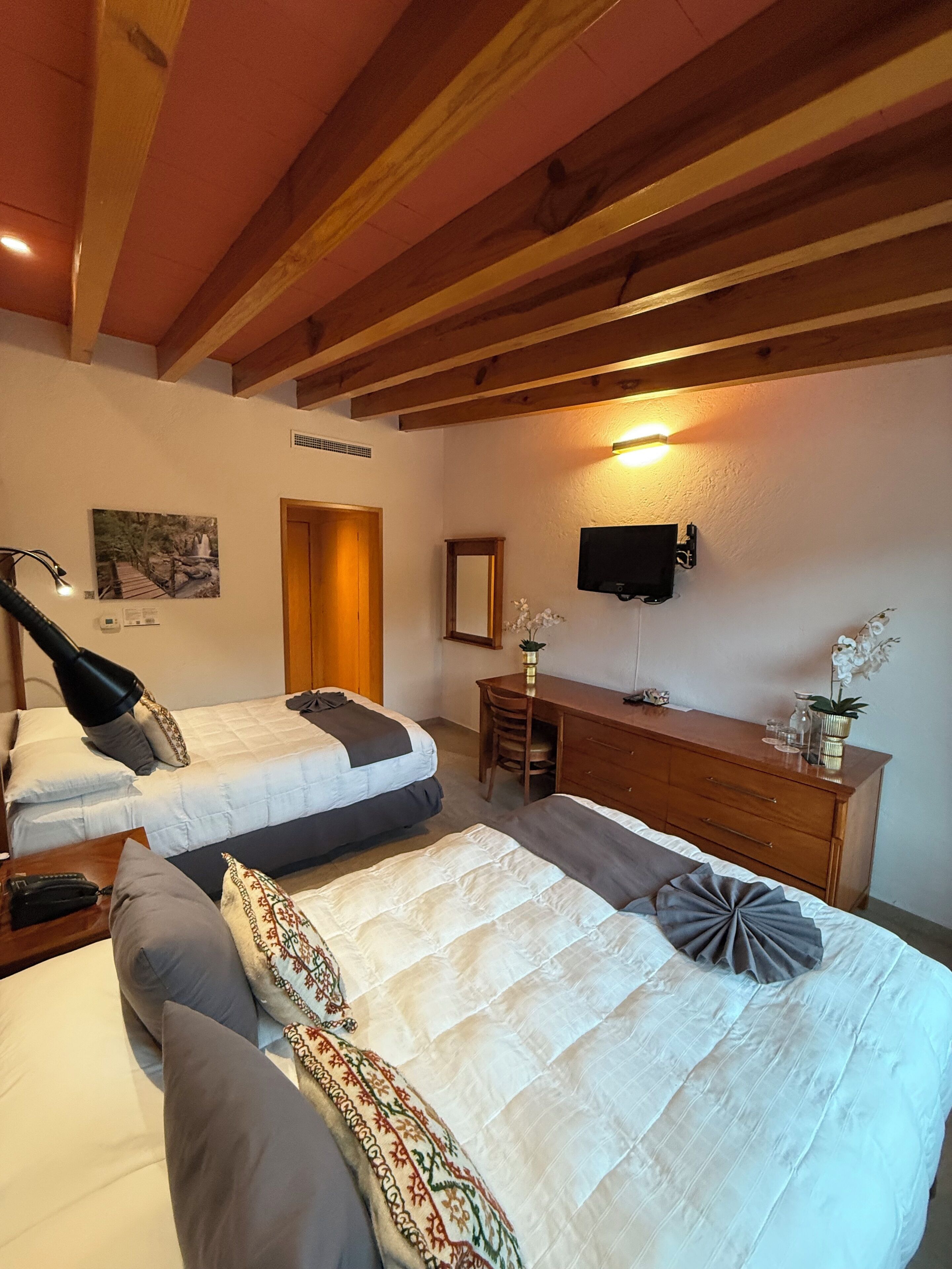 A hotel room with two beds, a wooden ceiling, a desk with a chair, a TV, and a wall-mounted mirror.