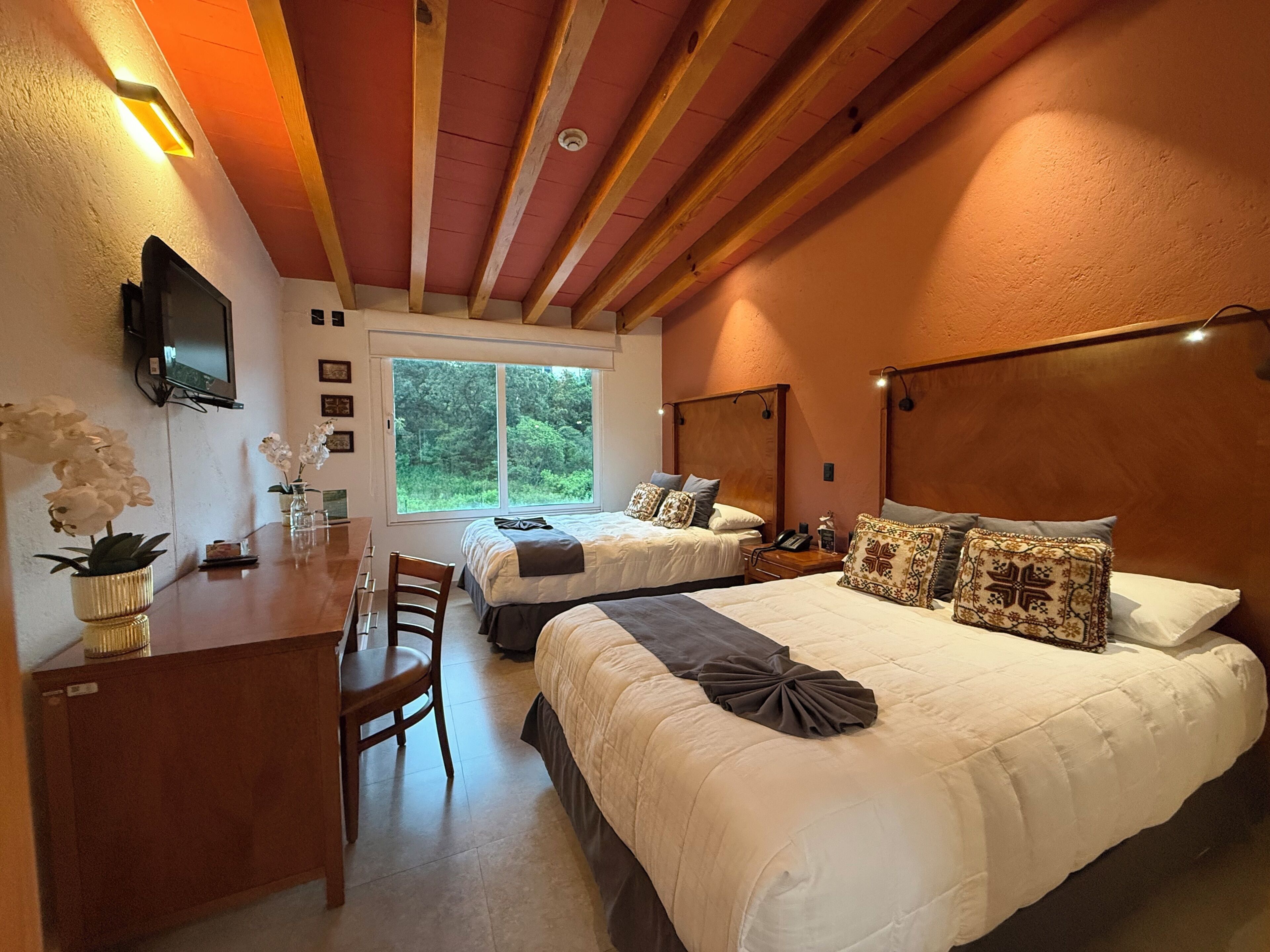 Habitación de hotel con techo de madera, dos camas con almohadones decorativos, un escritorio con silla, un televisor montado en la pared y una ventana con vista a follaje.