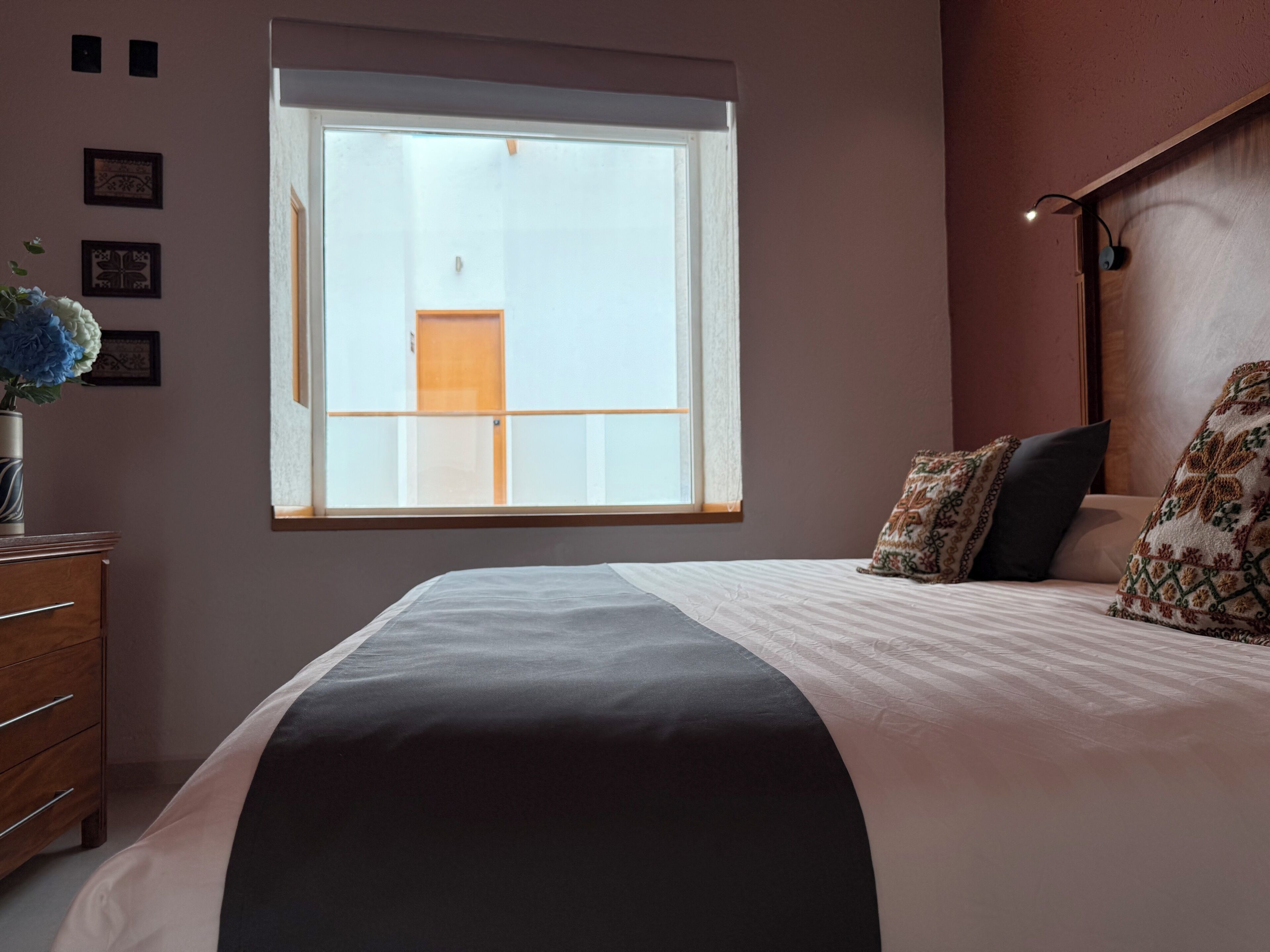A bedroom with a large window, a bed with patterned pillows, a bedside table, and a dresser.