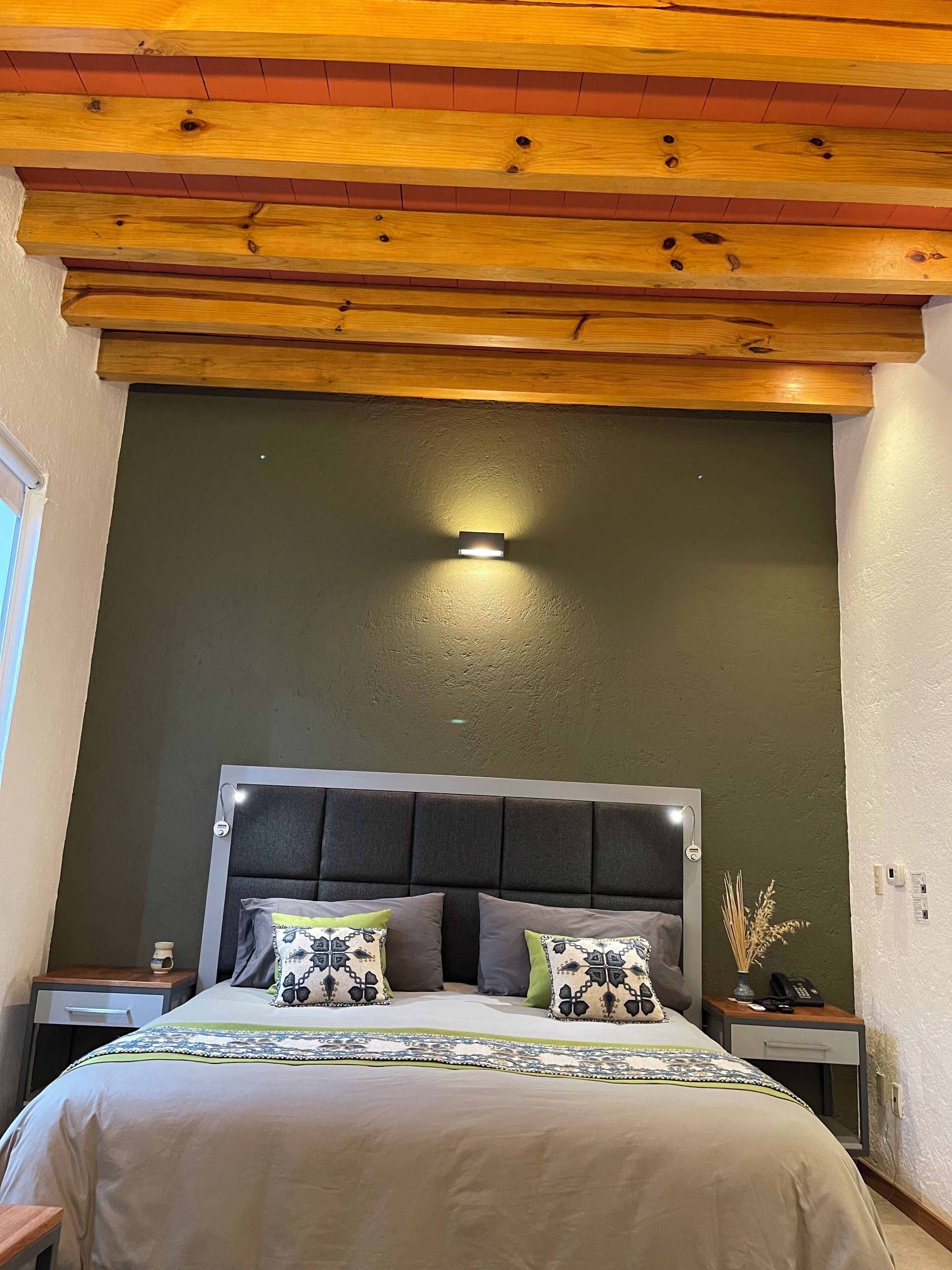 A bedroom with a bed, nightstands, a wall-mounted light, and a wooden ceiling.