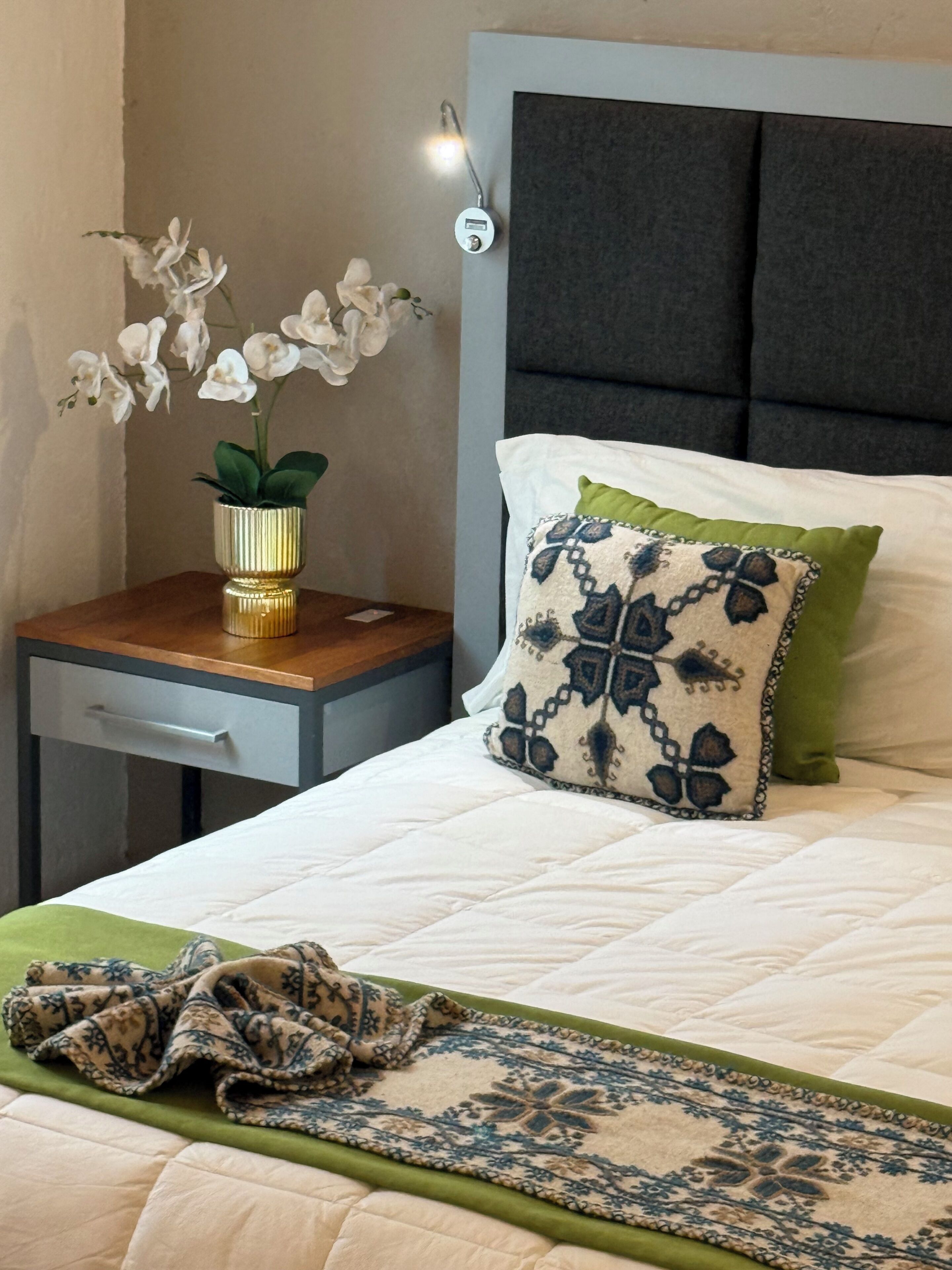Una cama bien tendida con almohadas estampadas, una mesita de noche con un florero de flores blancas, y una lámpara de pared.