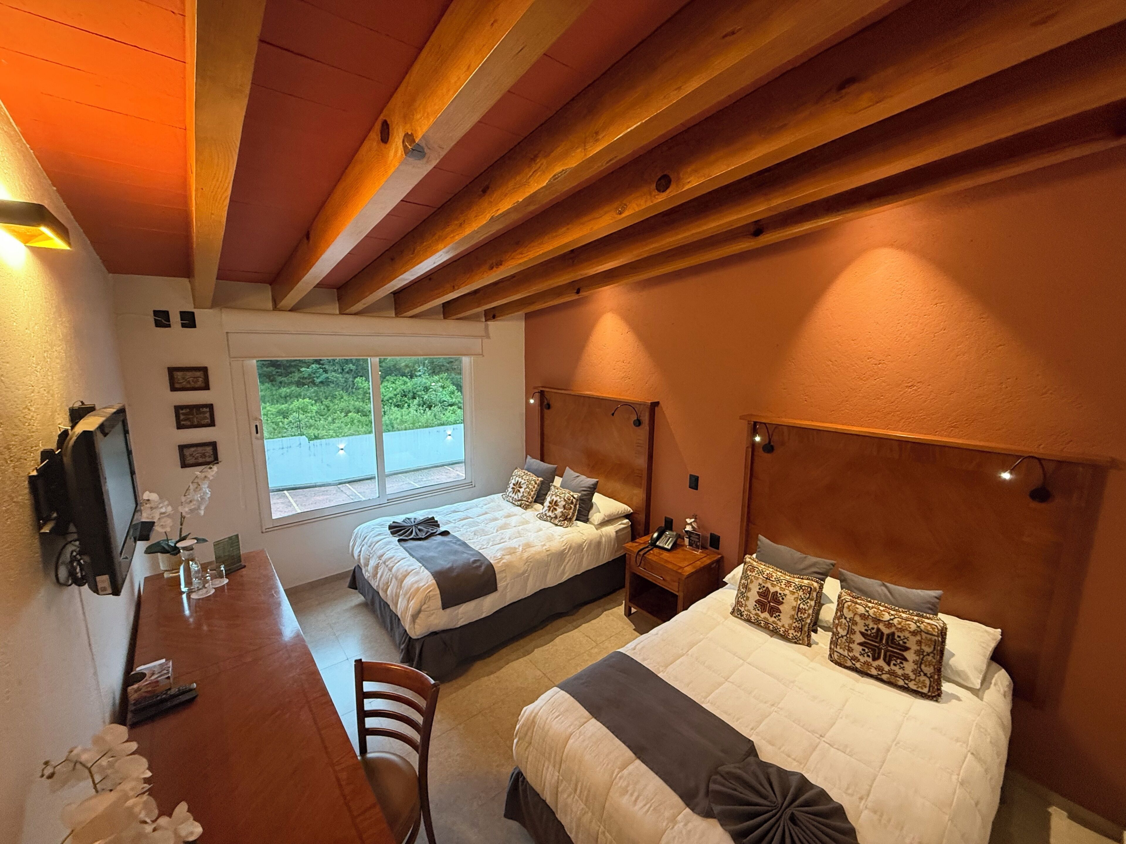 A bedroom with two beds, a wooden ceiling, a desk with a chair, and a window with a view of greenery.