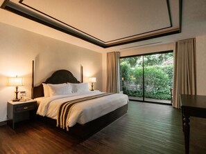 A hotel room with a large bed, two bedside tables with lamps, a wooden floor, and a view of greenery through a sliding glass door. - Burirattana (Chiang Mai)