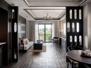 A modern hotel room with a wooden floor, a sofa with cushions, a dining table with chairs, and a balcony with a view of buildings and trees. - Burirattana (Chiang Mai)