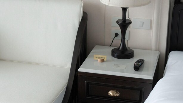 A hotel room with a white upholstered sofa, a bedside table with a lamp, a remote control, and a 'No Smoking' sign. - Burirattana (Chiang Mai)