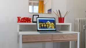 Ein Laptop steht auf einem weißen Schreibtisch, darüber ein Spiegel, ein Bilderrahmen und eine rote Tasse mit Stöckchen.