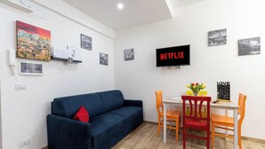 Ein Wohnzimmer mit blauem Sofa, einem Esstisch mit Stühlen, einem an der Wand montierten Fernseher und mehreren gerahmten Bildern.