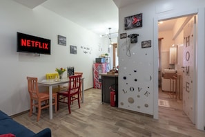 Una sala moderna con una televisión mostrando 'NETFLIX', un comedor con sillas y una mesa, y una cocina con un refrigerador y una cafetera.