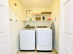 A laundry room with two front-loading washing machines, a shelf with various items, and a towel rack.