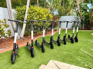 A row of stationary scooters parked on a green lawn with a white wall and tropical plants in the background.