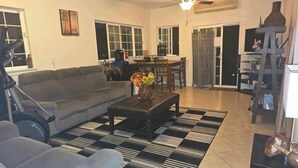 A living room with a sofa, coffee table, and a patterned rug. There is a ceiling fan, a dining area with chairs, and a kitchen in the background. - Robbins Estate (Wailuku)