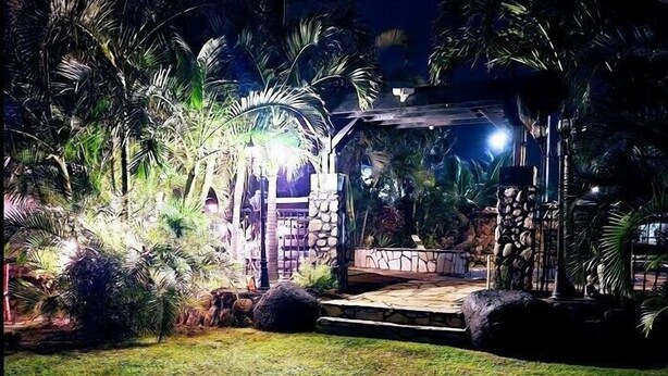 A well-lit outdoor area with a stone structure, possibly a gazebo, surrounded by lush greenery and palm trees. - Robbins Estate (Wailuku)