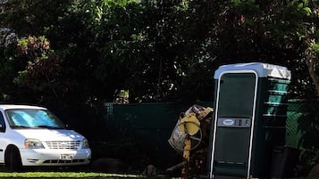 A white car parked next to a portable toilet in a grassy area with trees in the background.
