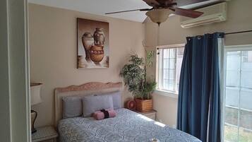 A bedroom with a bed, a ceiling fan, a window with curtains, a wall-mounted air conditioner, a potted plant, and a painting.