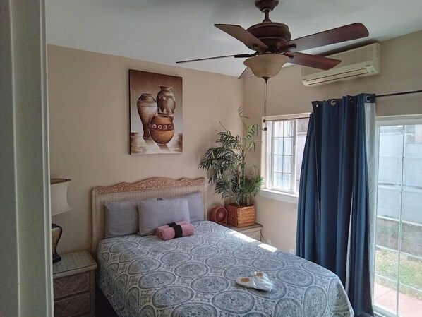 A bedroom with a bed, a ceiling fan, a window with curtains, a wall-mounted air conditioner, a potted plant, and a painting. - Robbins Estate (Wailuku)